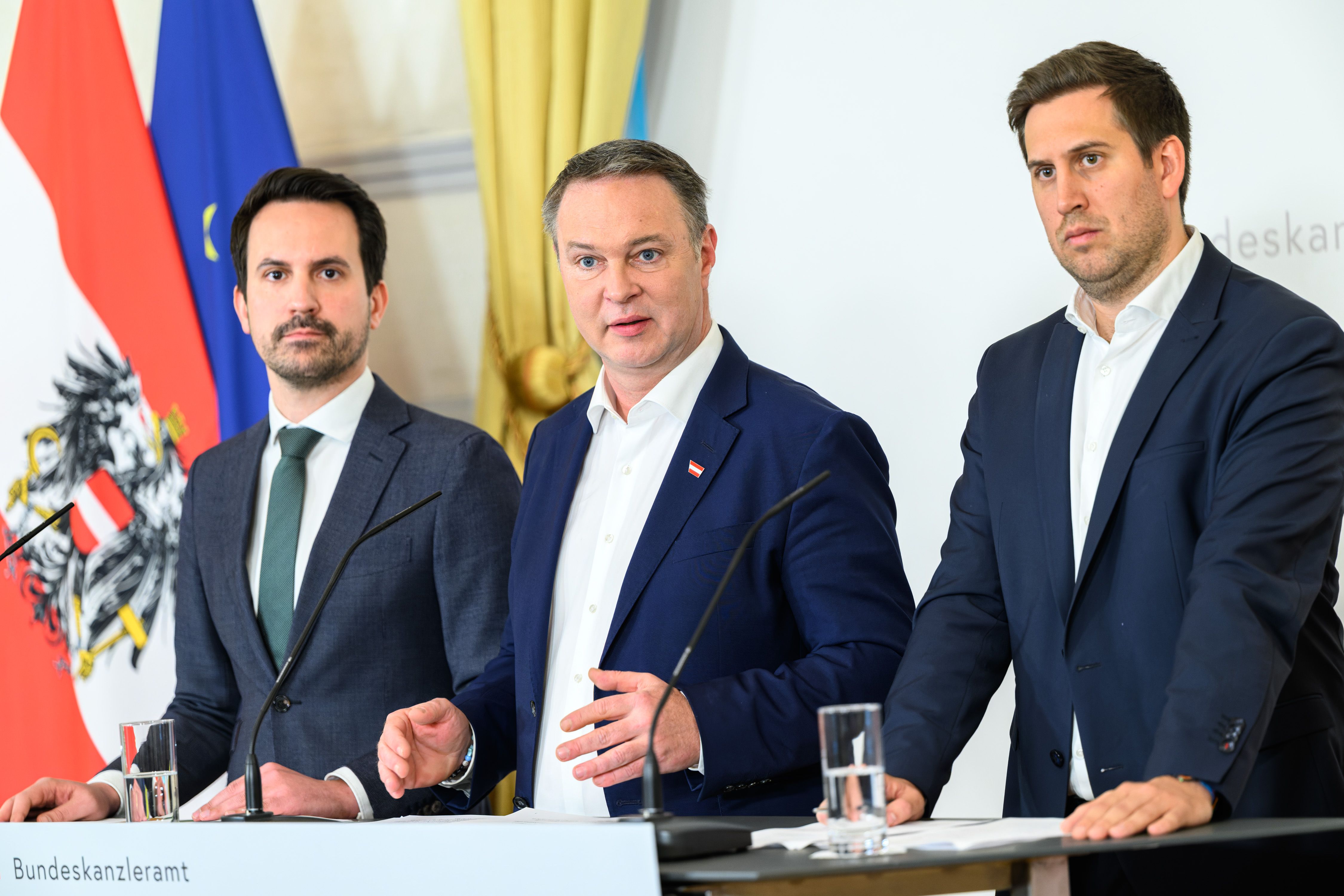 Drei Männer im Anzug sprechen bei einer Pressekonferenz in Wien vor Mikrofonen und Fahnen.