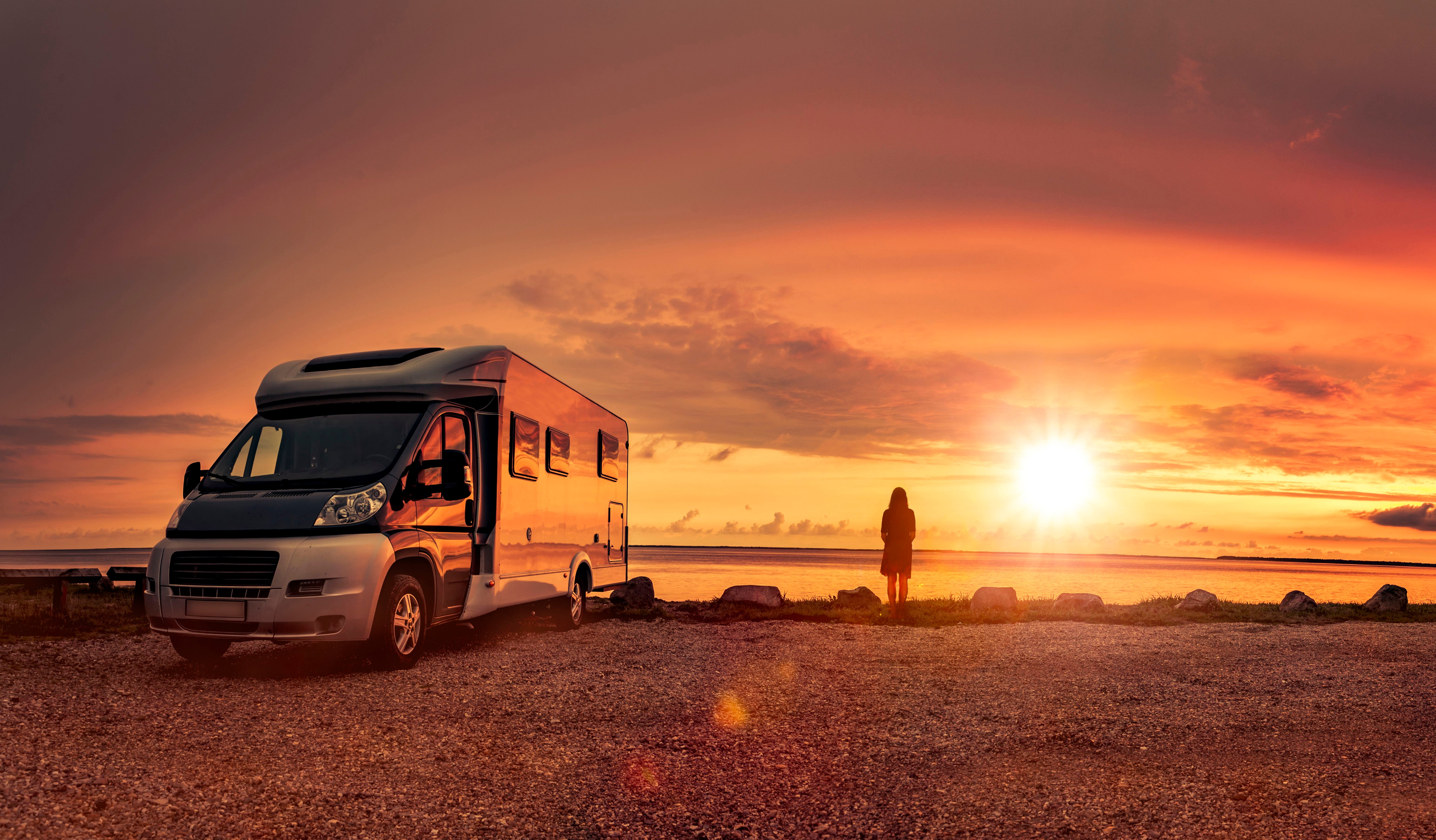 Frau steht bei Sonnenuntergang am Strand neben einem Wohnmobil.