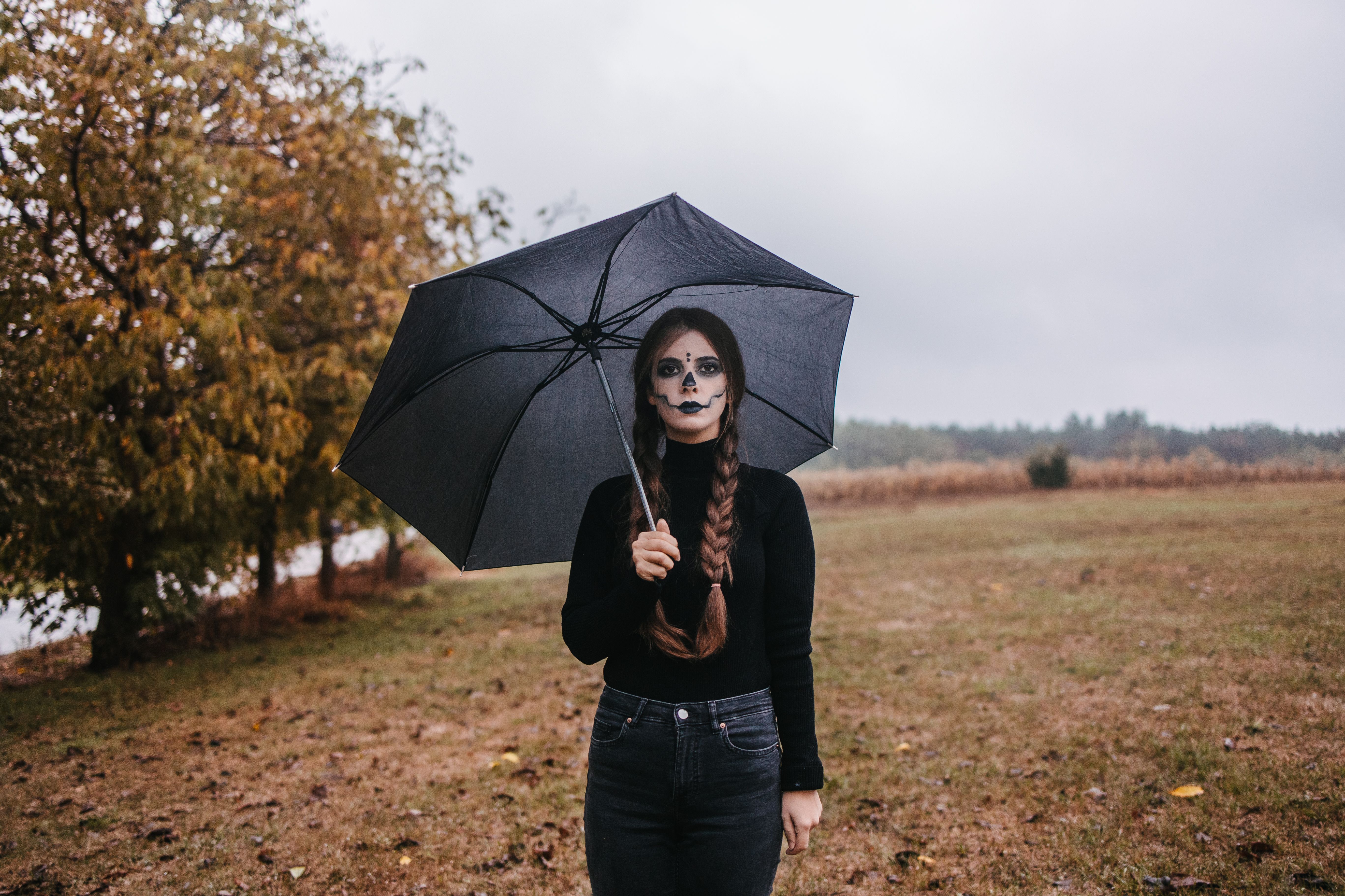 Frau mit Zöpfen und Skelett-Make-up hält schwarzen Regenschirm auf einem trüben Feld.