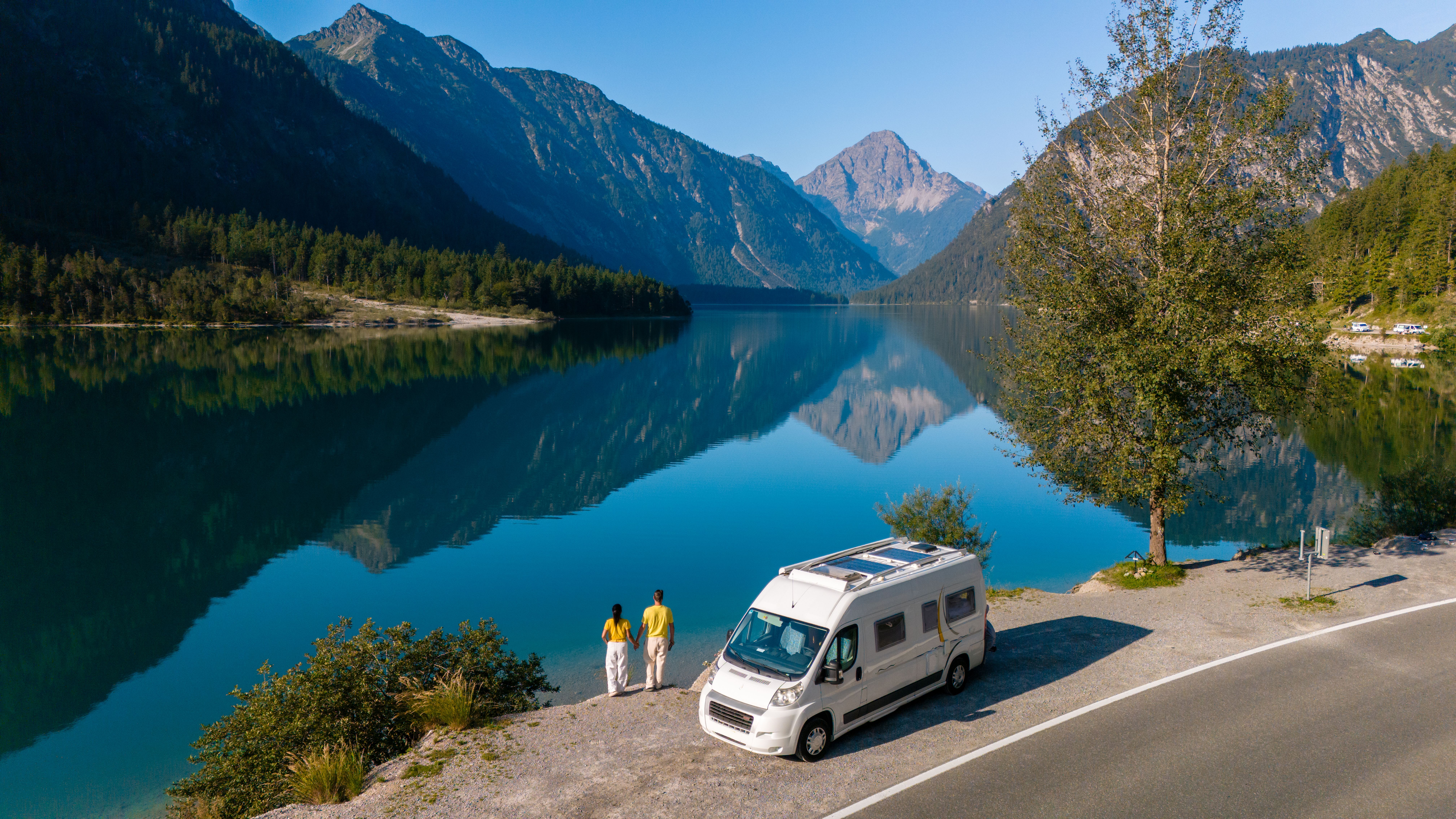 Zwei Reisende stehen am Plansee vor einem Wohnmobil und betrachten die Spiegelung der Berge im Wasser.