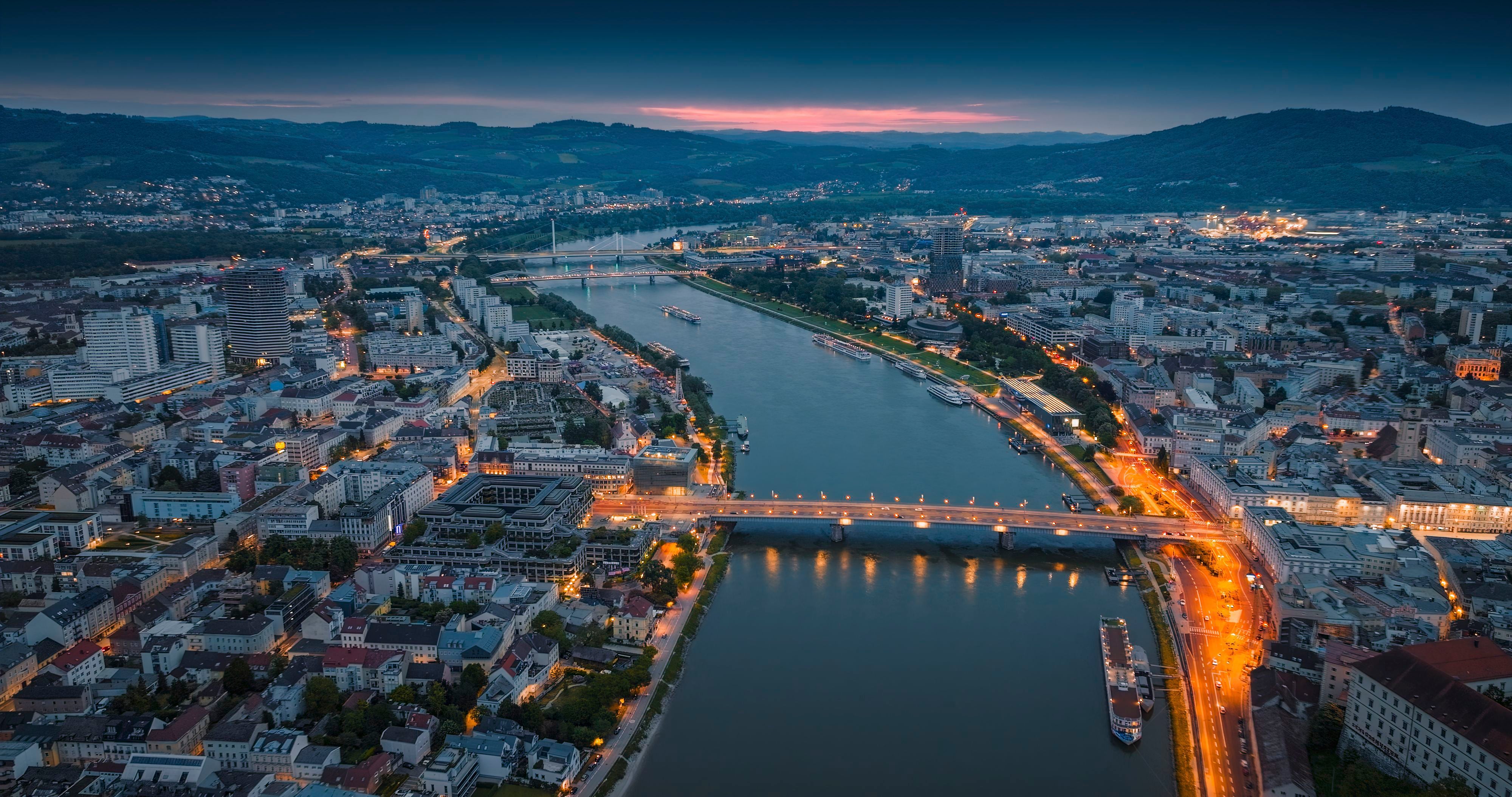 Luftaufnahme von Linz bei Dämmerung mit beleuchteten Brücken über die Donau und umliegenden Hügeln.