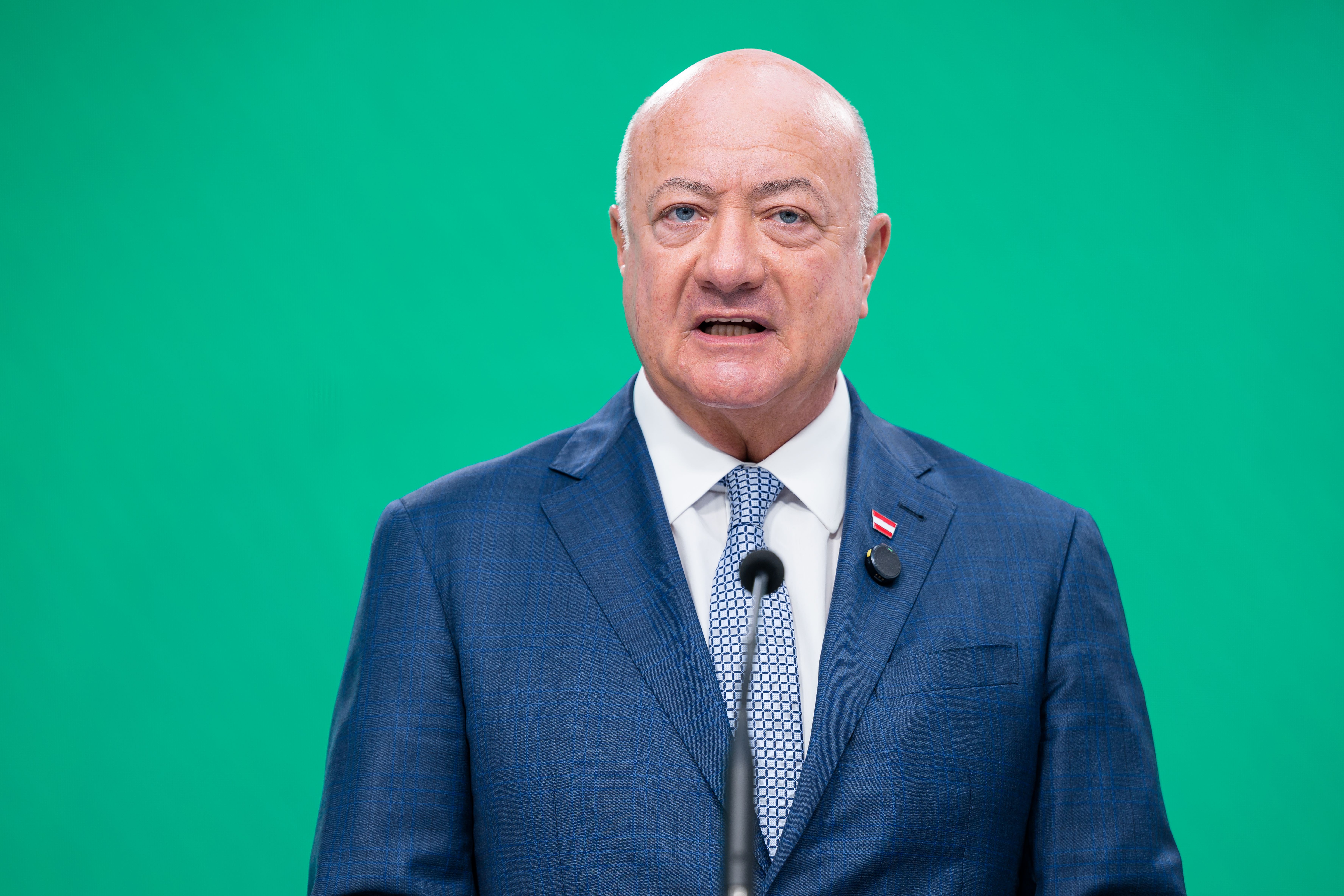 Ein Mann im blauen Anzug spricht bei einer Pressekonferenz vor grünem Hintergrund.