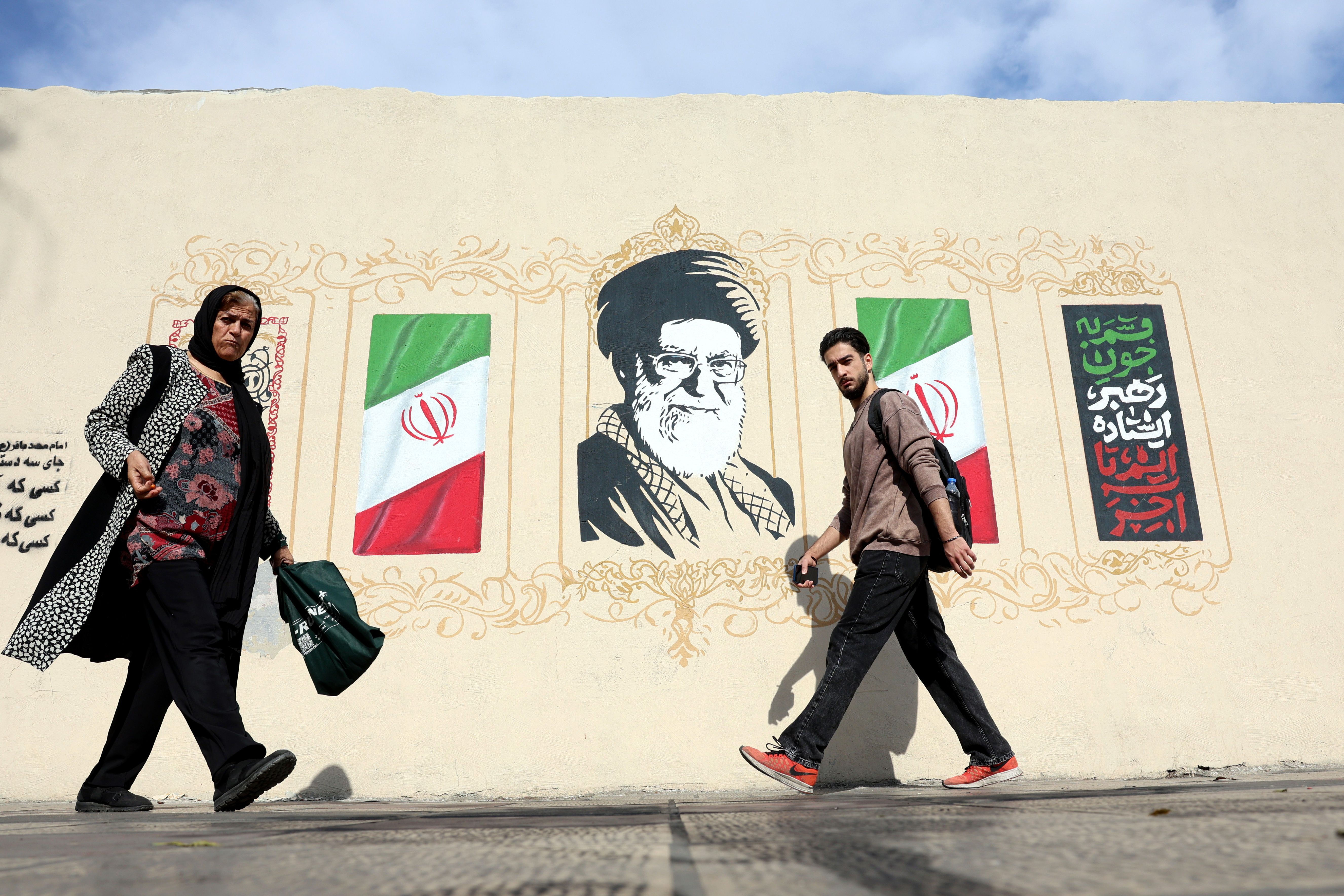 Zwei Menschen gehen an einem Wandbild mit iranischen Flaggen und einem Porträt vorbei.