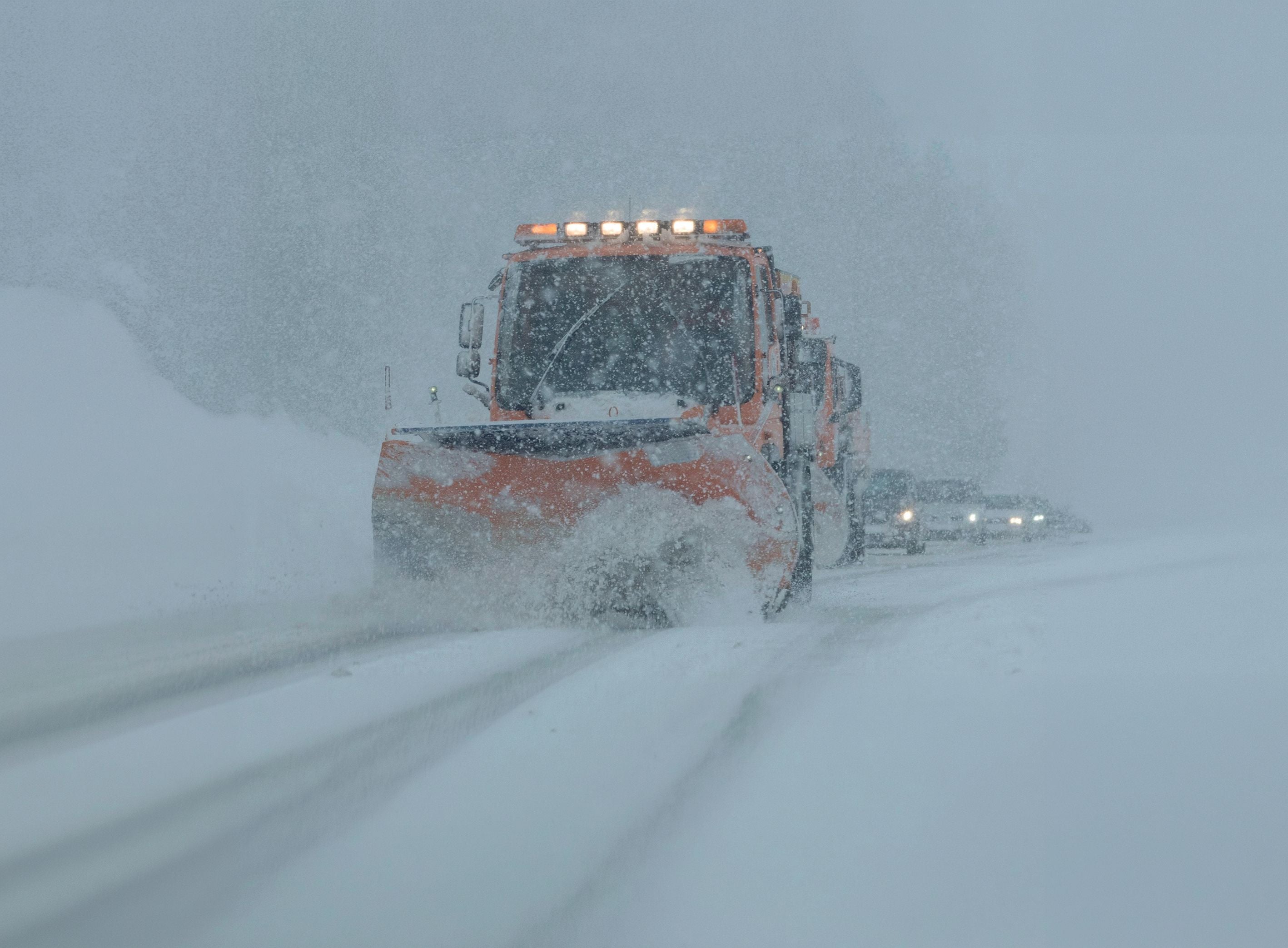 Schneepflug räumt bei starkem Schneefall eine verschneite Straße, Autos folgen dahinter.