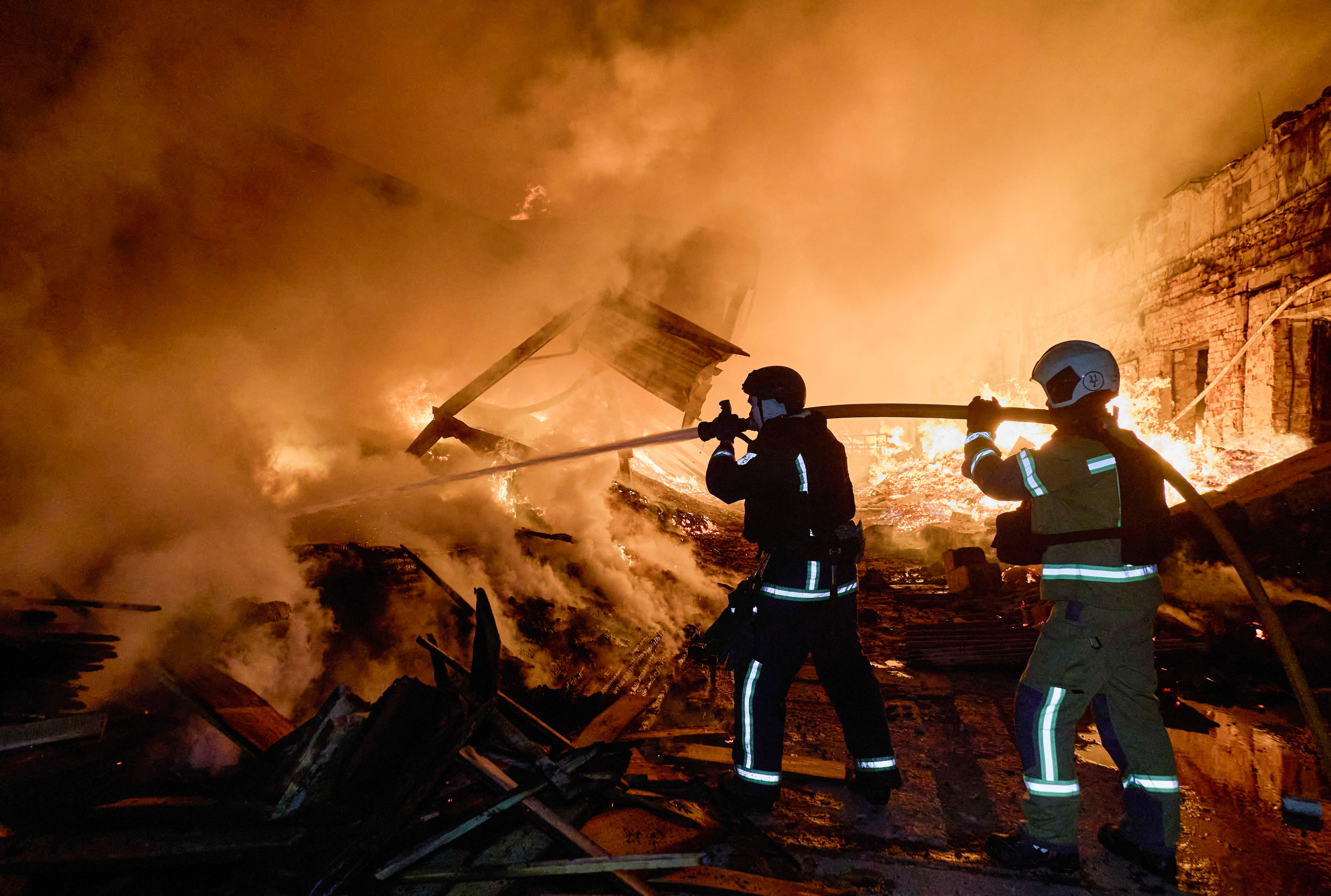 Zwei Feuerwehrleute löschen nachts einen Großbrand mit starkem Rauch und Flammen.
